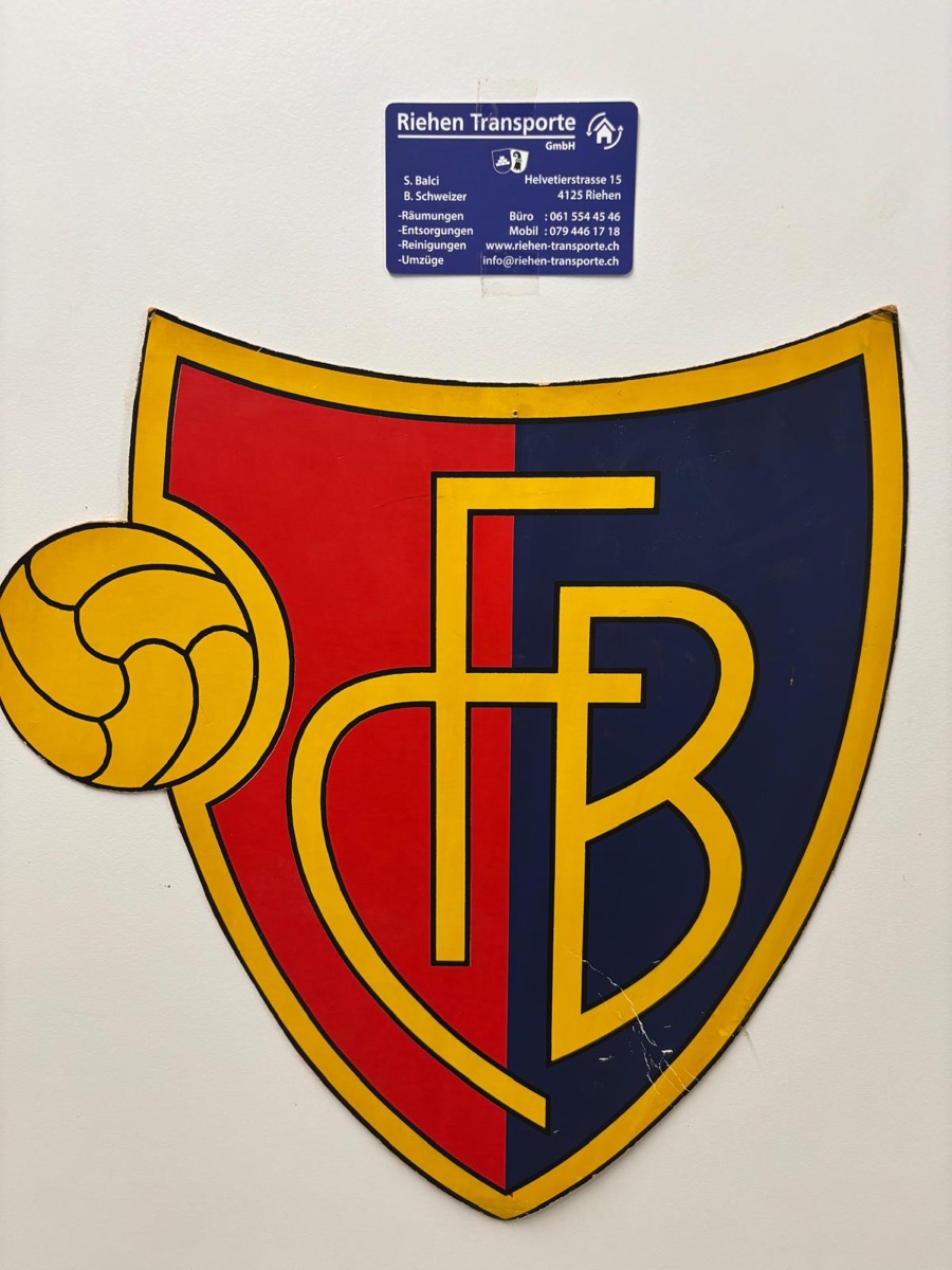Aufkleber der Riehen Transporte GmbH neben dem rot-blauen FCB-Wappen an der Bürowand